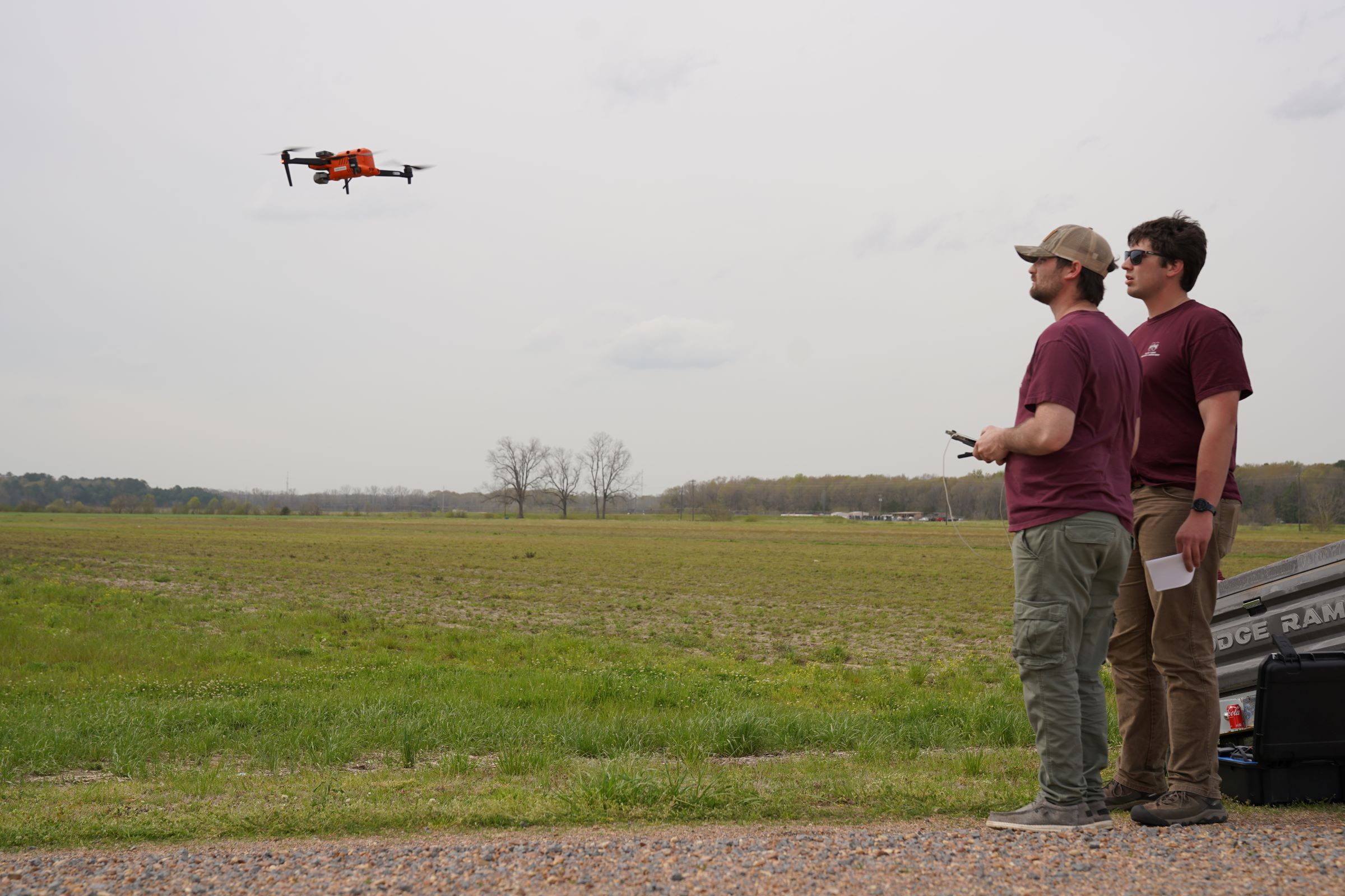 Two people, including a drone pilot, watch as a drone hovers nearby.  